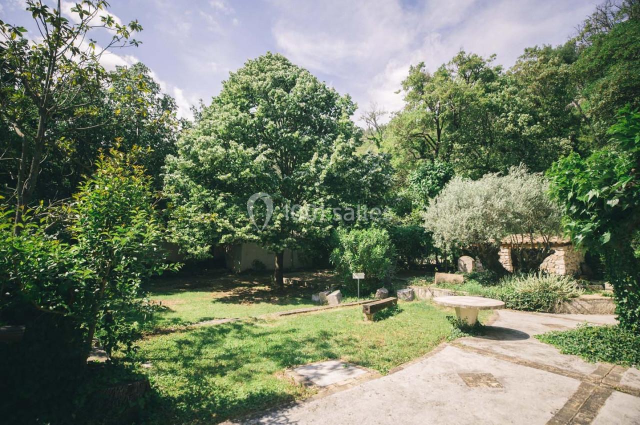 Jardin arboré avec des arbres, des buissons et une table en pierre sous un ciel partiellement nuageux.