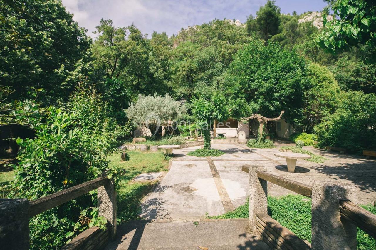 Jardin verdoyant avec des arbres, des tables en pierre et un chemin pavé, vu depuis un escalier en bois.