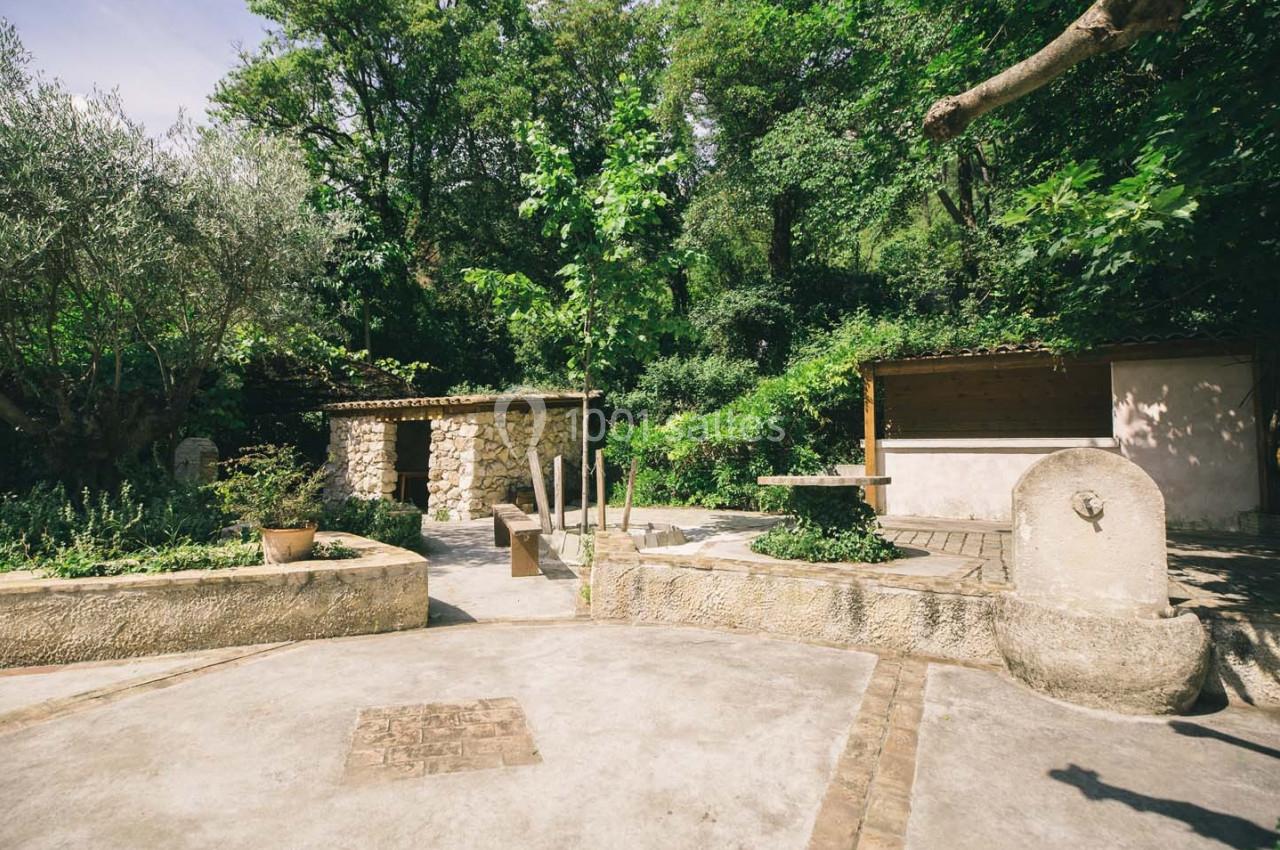 Cour extérieure ensoleillée avec fontaine, bancs en pierre, végétation dense et petites structures en pierre et bois.