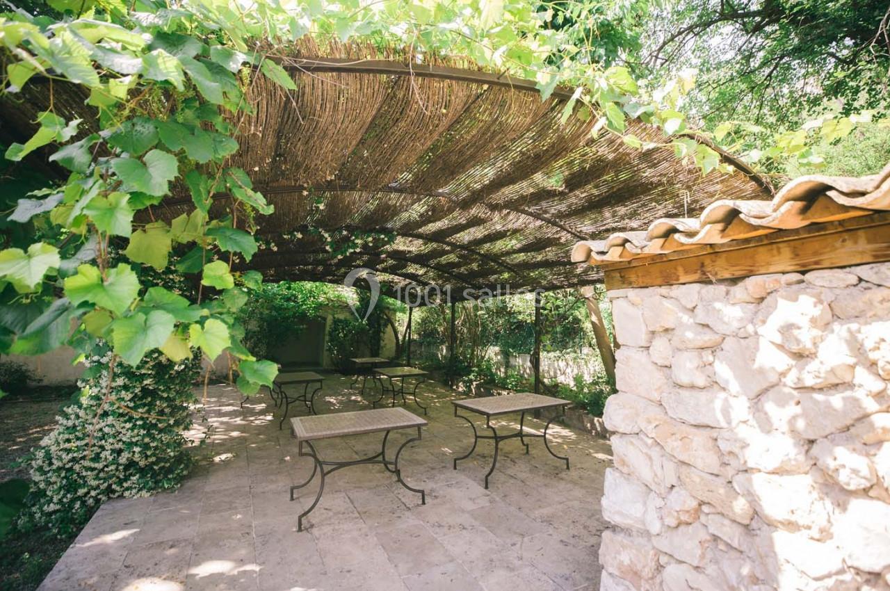 Terrasse ombragée avec pergola en bois et feuillage, tables en métal et mur en pierre dans un cadre naturel.