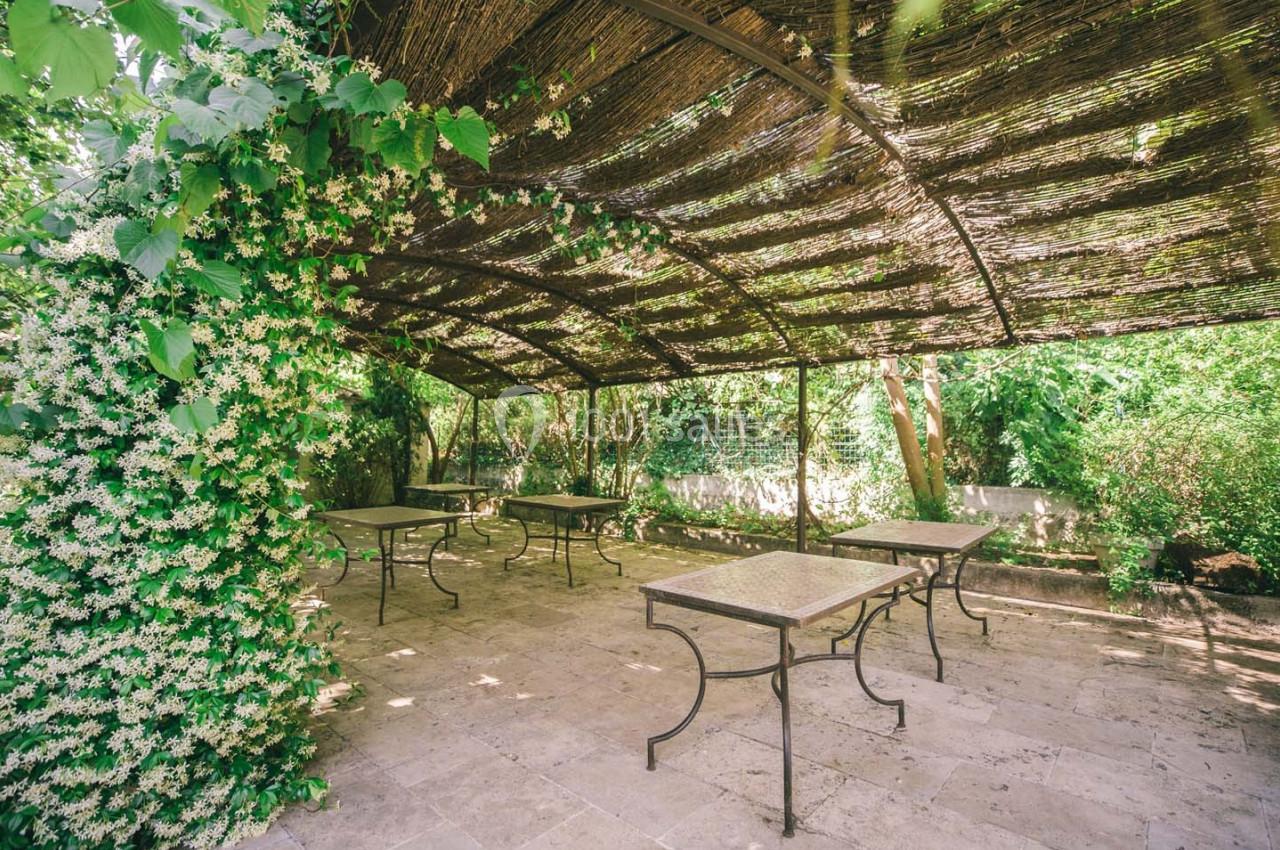 Terrasse ombragée avec pergola en bois, tables en métal et plantes grimpantes dans un jardin verdoyant.