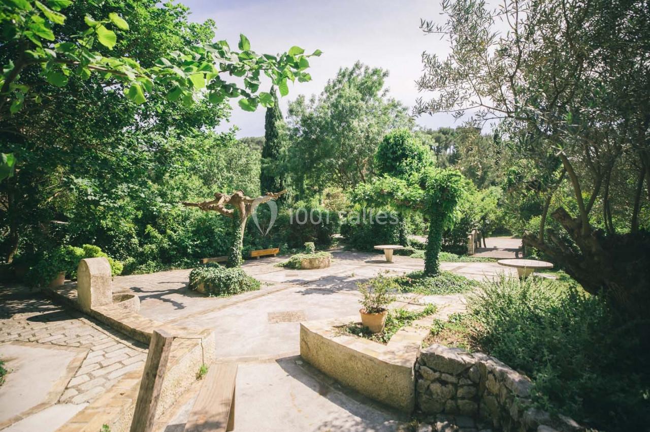 Jardin arboré avec allées pavées, bancs en bois et végétation méditerranéenne sous un ciel dégagé.