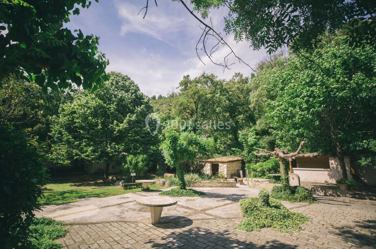 Cour extérieure pavée entourée de végétation dense, avec tables en pierre et petite structure en arrière-plan.