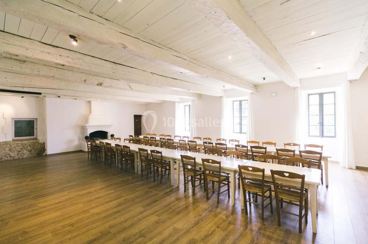 Grande salle lumineuse avec parquet, poutres apparentes blanches, tables alignées et chaises en bois.
