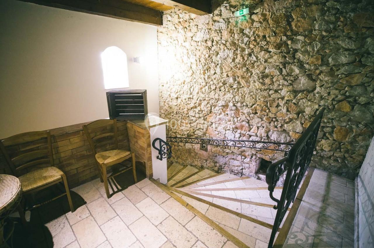 Escalier en pierre avec mur en pierre apparente, éclairé par une petite fenêtre, et chaises en bois à gauche.