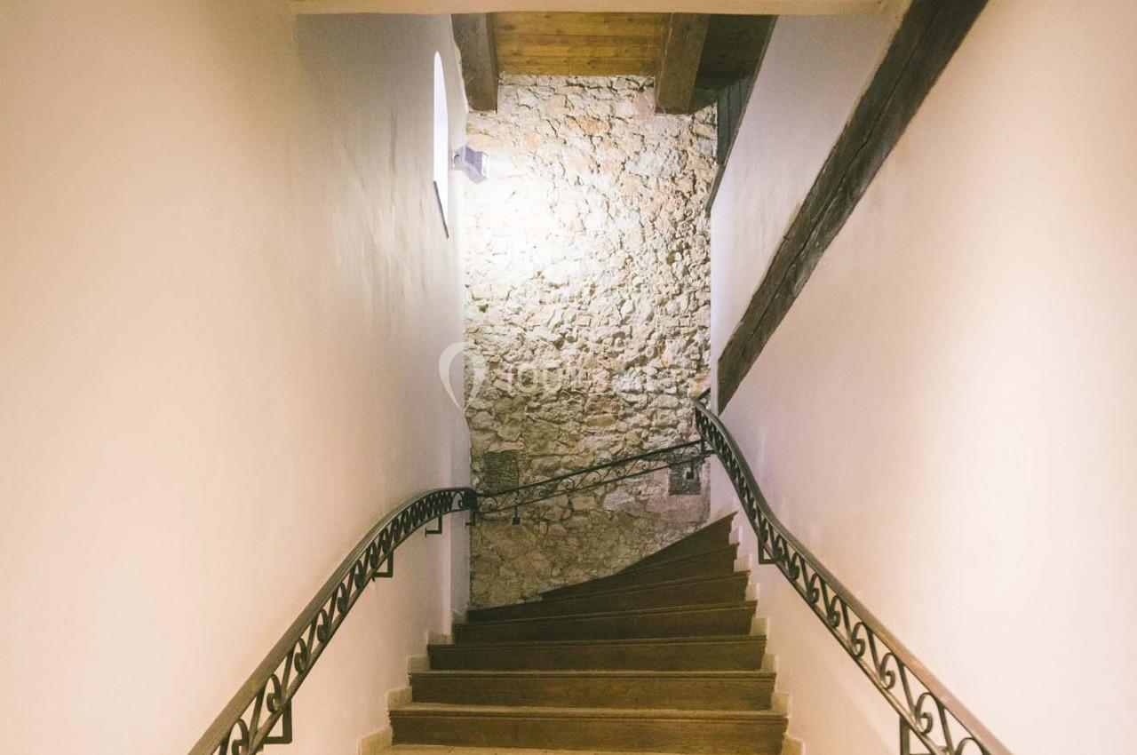 Escalier en bois descendant avec des murs en pierre et des rampes en fer forgé, éclairé par une lumière murale.