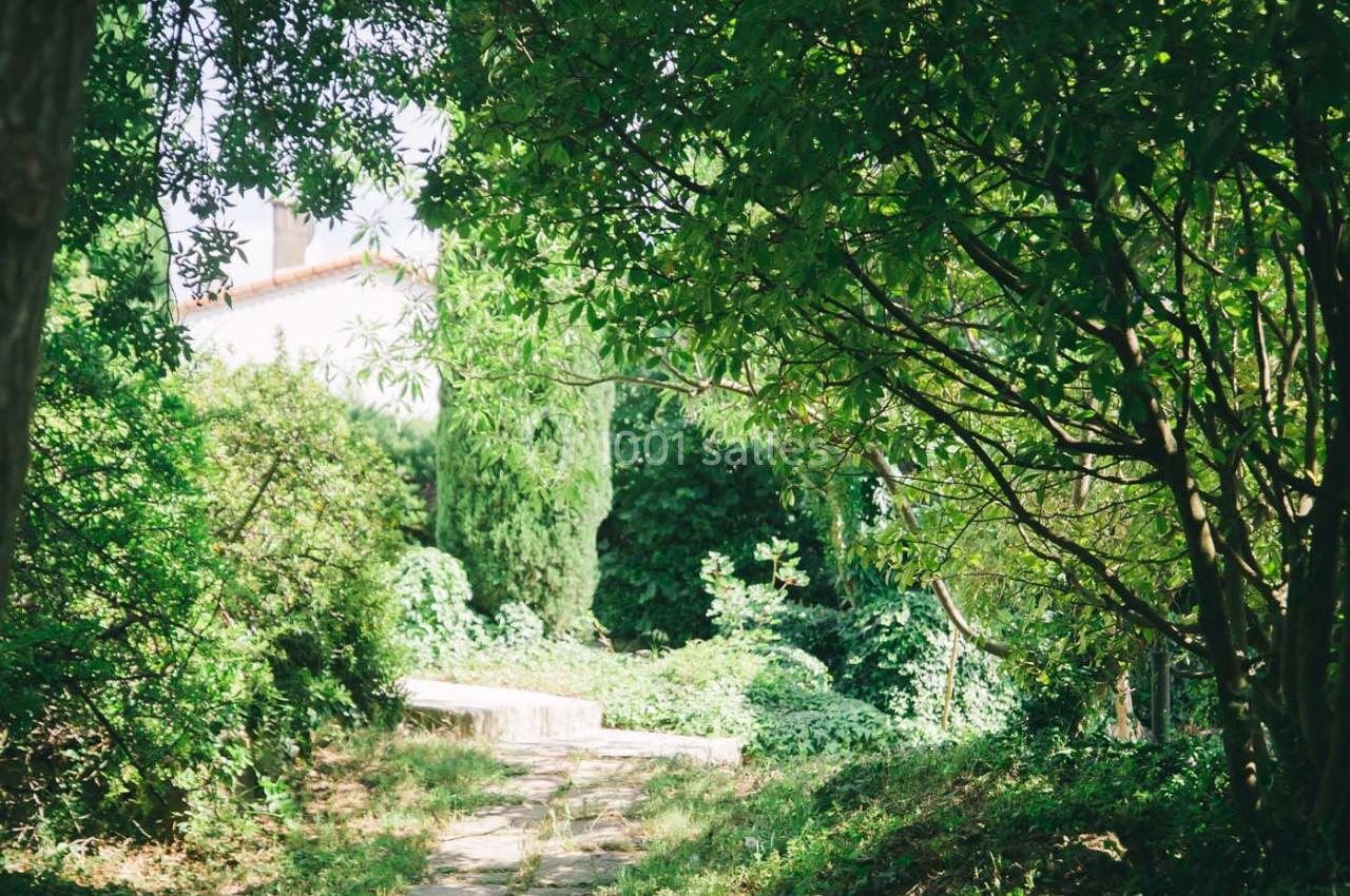 Chemin en pierre bordé de végétation dense menant à une maison blanche partiellement visible à travers les arbres.