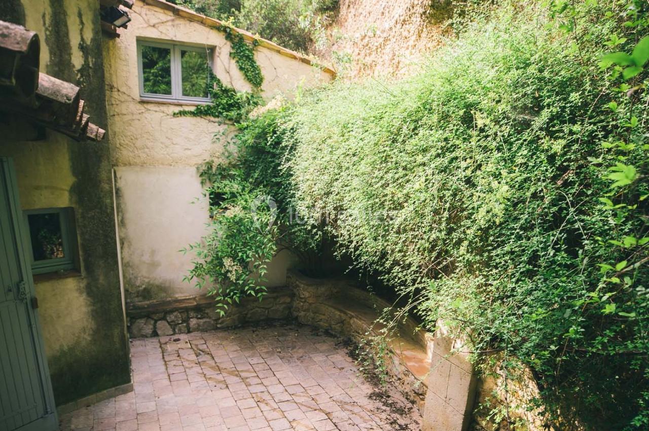 Cour intérieure pavée entourée de murs en pierre, avec des plantes grimpantes et des fenêtres sur une façade.