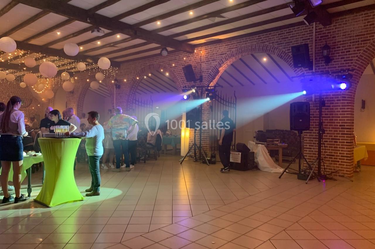 Salle de réception éclairée avec des guirlandes lumineuses, des invités debout près de tables hautes et une ambiance festive.