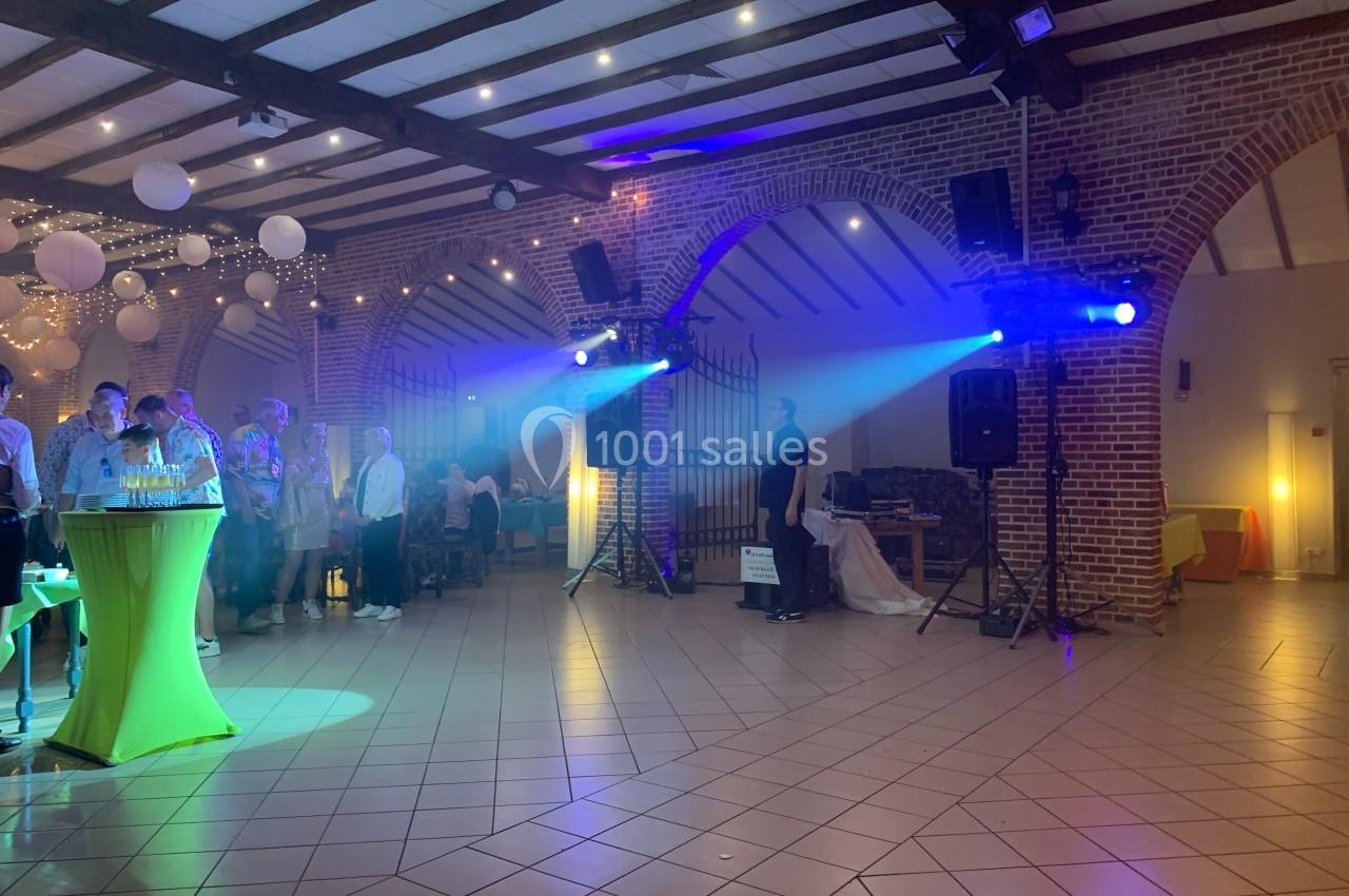 Salle de réception avec invités debout et assis, éclairages colorés et décorations suspendues sous un plafond en poutres…