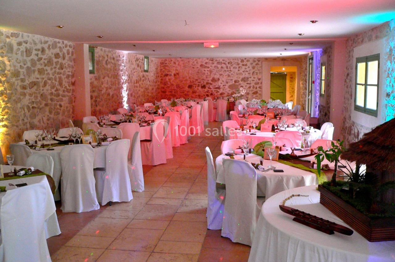 Salle de réception en pierre avec tables rondes dressées, nappes blanches et éclairage coloré.