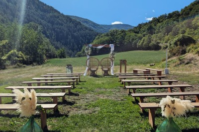 Un homme en costume parle devant une arche décorée de fleurs séchées, dans un cadre naturel avec des montagnes et des arbres.