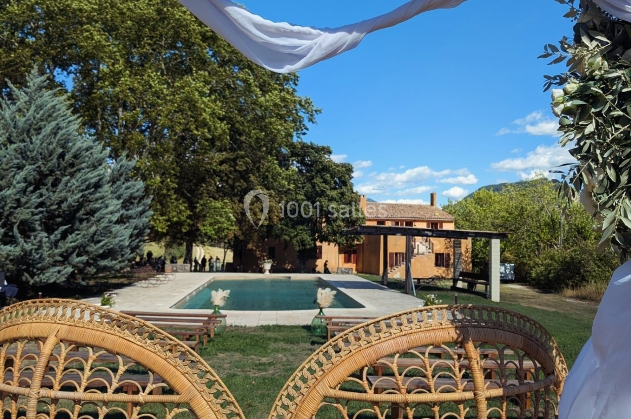 Vue sur une piscine entourée de verdure et de chaises en rotin, avec une maison en arrière-plan sous un ciel bleu.