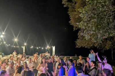 Un homme en costume parle devant une arche décorée de fleurs séchées, dans un cadre naturel avec des montagnes et des arbres.