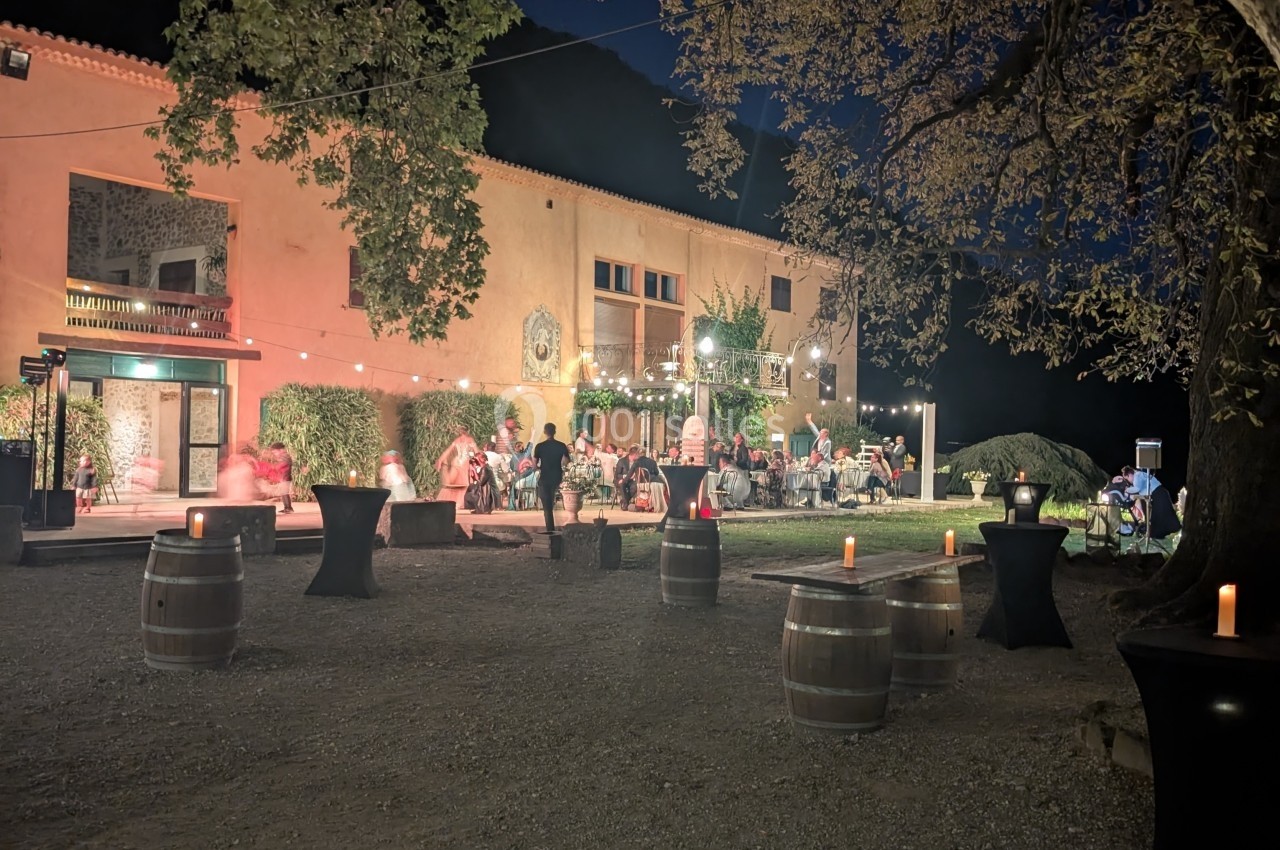 Soirée en plein air devant un bâtiment éclairé, avec des invités assis et des tonneaux utilisés comme tables.