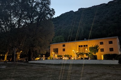 Un homme en costume parle devant une arche décorée de fleurs séchées, dans un cadre naturel avec des montagnes et des arbres.