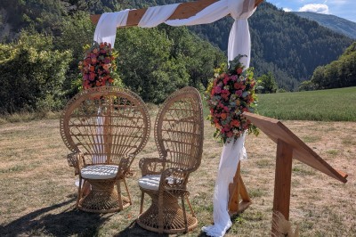 Un homme en costume parle devant une arche décorée de fleurs séchées, dans un cadre naturel avec des montagnes et des arbres.