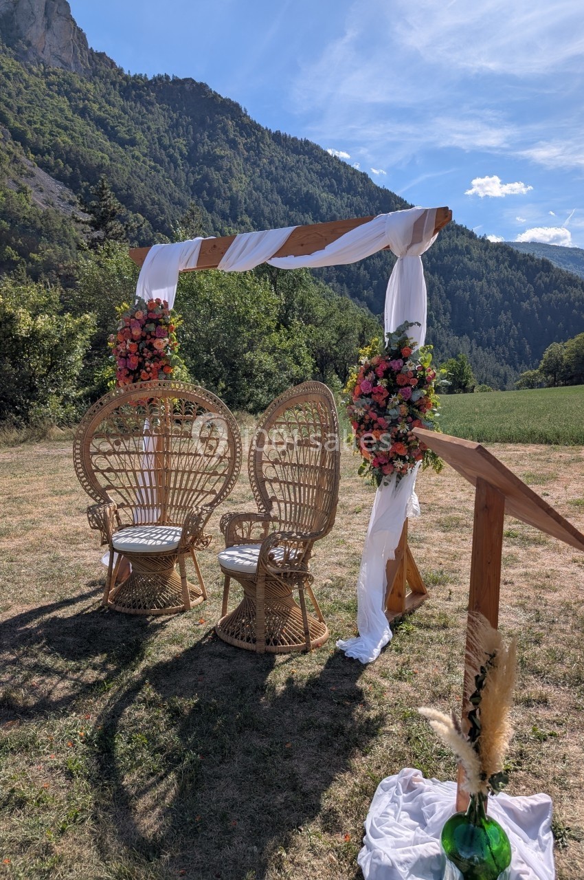 Arche de mariage décorée de fleurs et de tissu blanc, avec deux fauteuils en osier, dans un cadre naturel montagneux.