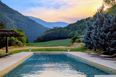 Un homme en costume parle devant une arche décorée de fleurs séchées, dans un cadre naturel avec des montagnes et des arbres.