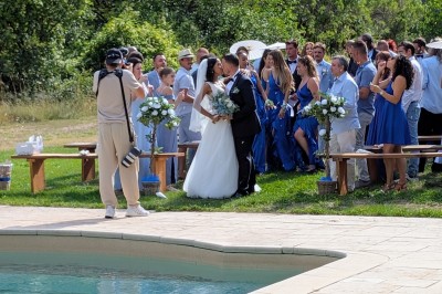 Un homme en costume parle devant une arche décorée de fleurs séchées, dans un cadre naturel avec des montagnes et des arbres.