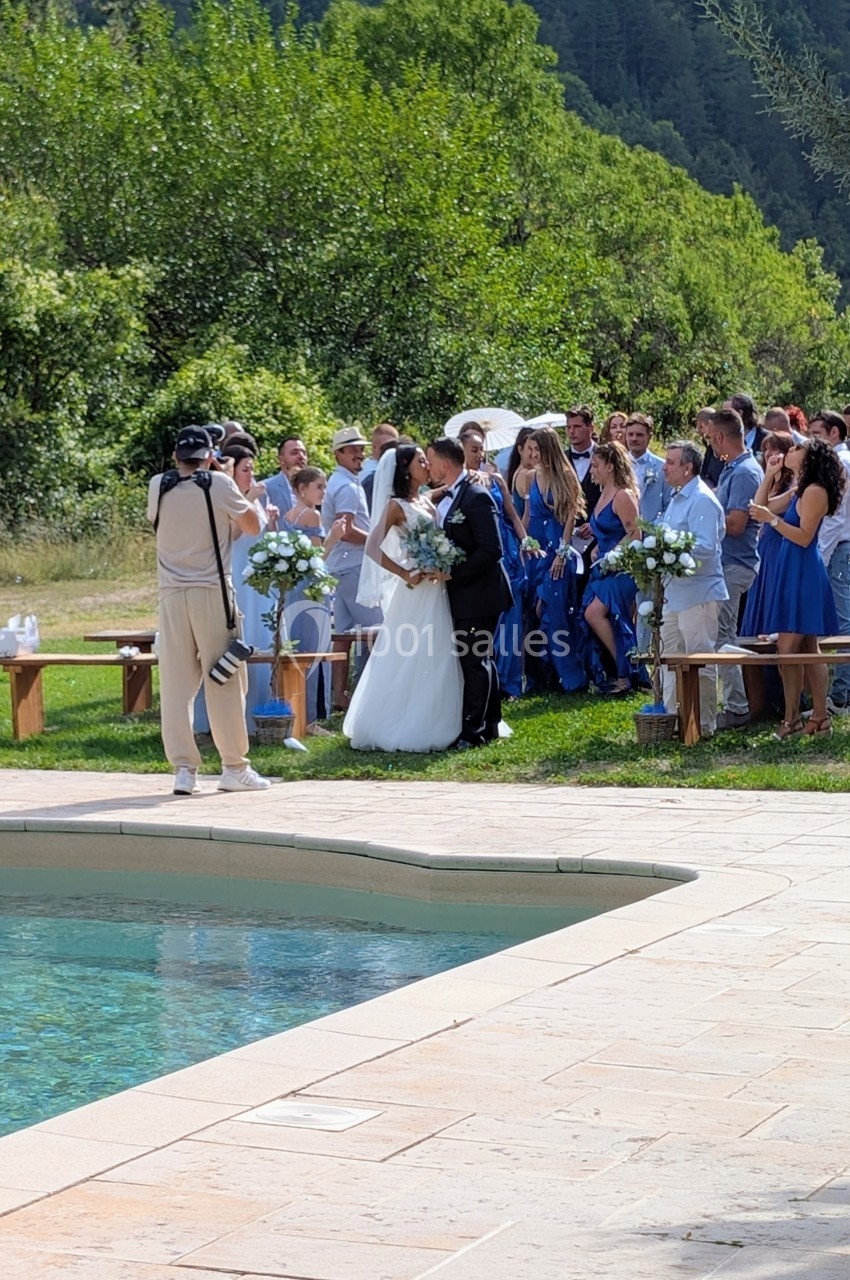Un couple de mariés pose devant des invités en tenue élégante près d'une piscine, dans un cadre naturel.
