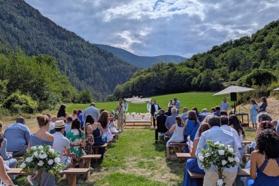 Un homme en costume parle devant une arche décorée de fleurs séchées, dans un cadre naturel avec des montagnes et des arbres.