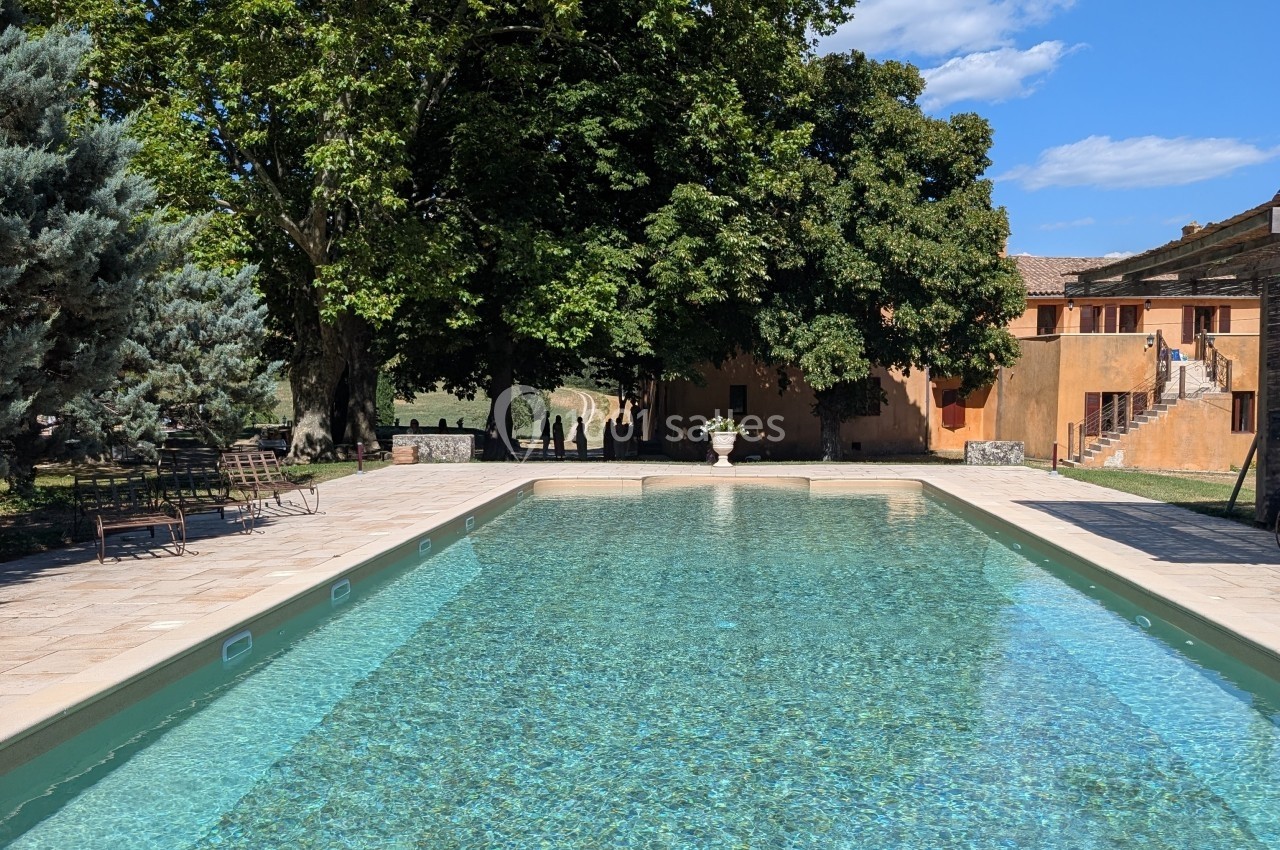 Piscine extérieure entourée de végétation et de bâtiments en pierre sous un ciel dégagé.