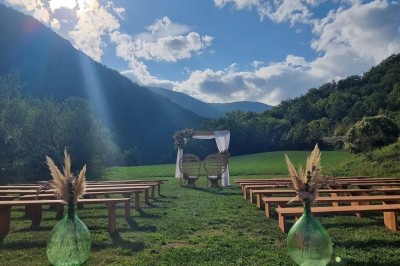 Un homme en costume parle devant une arche décorée de fleurs séchées, dans un cadre naturel avec des montagnes et des arbres.
