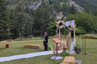 Un homme en costume parle devant une arche décorée de fleurs séchées, dans un cadre naturel avec des montagnes et des arbres.