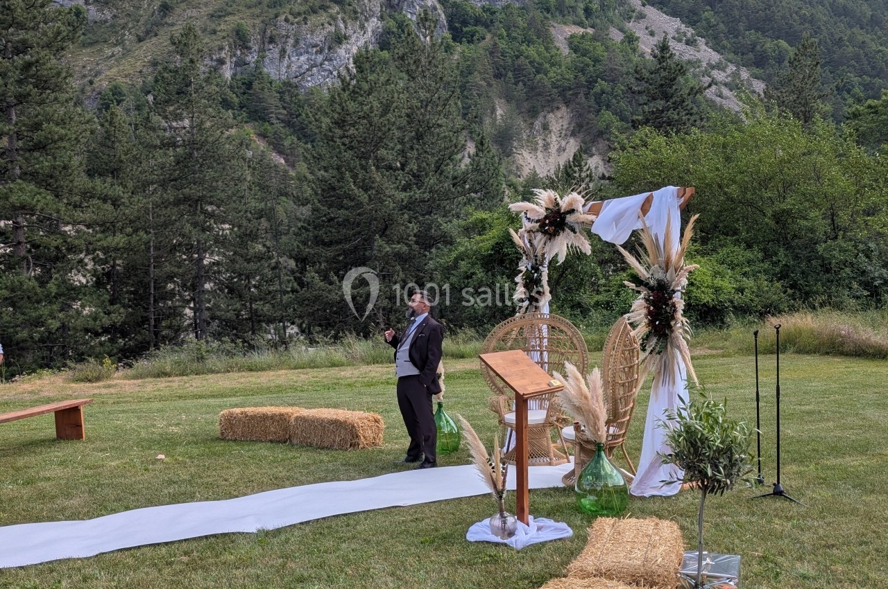 Un homme en costume parle devant une arche décorée de fleurs séchées, dans un cadre naturel avec des montagnes et des arbres.