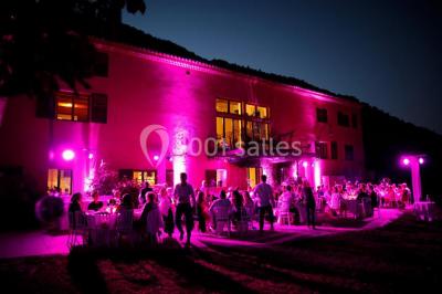 Un homme en costume parle devant une arche décorée de fleurs séchées, dans un cadre naturel avec des montagnes et des arbres.