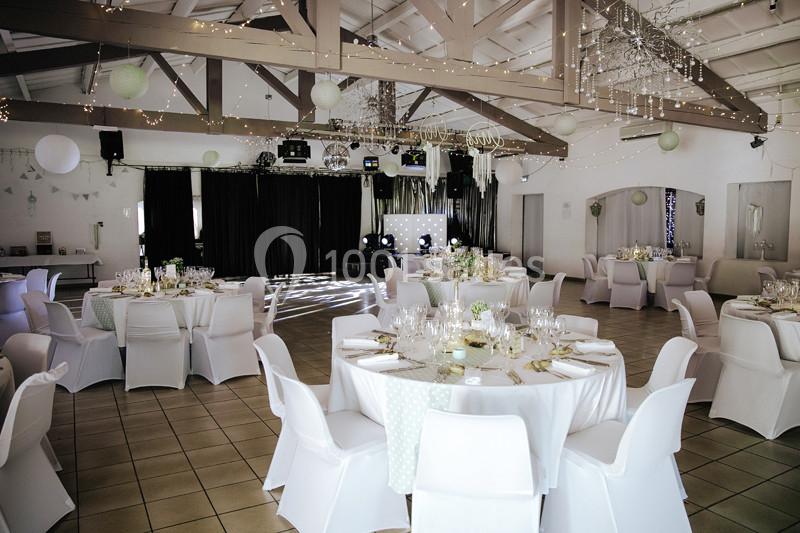 Salle de réception décorée avec des tables rondes dressées, des chaises blanches et des guirlandes lumineuses suspendues.