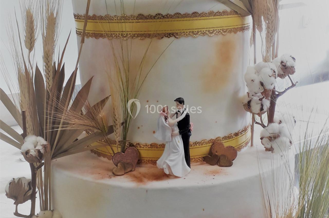 Gâteau de mariage à deux étages décoré de fleurs de coton, épis de blé et figurines de mariés.