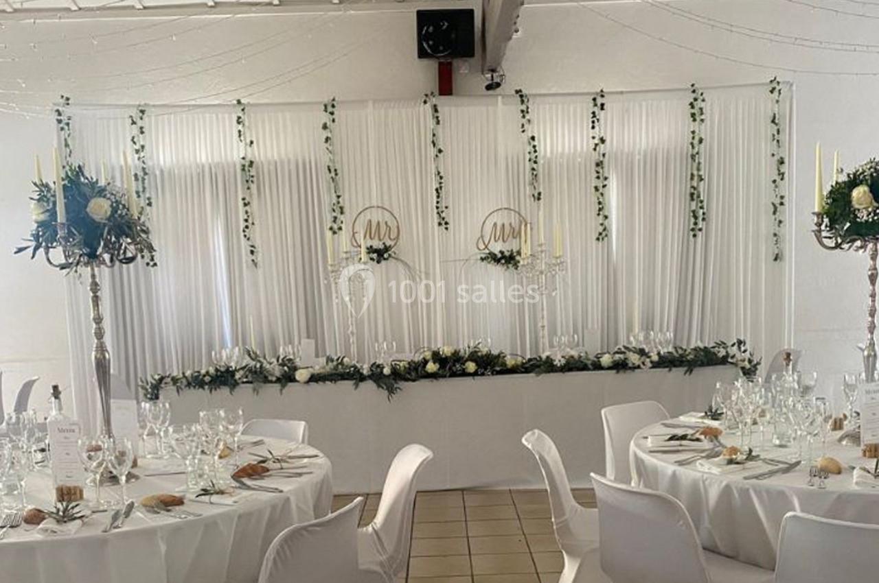 Salle de réception décorée avec des tables rondes, nappes blanches, chandeliers et une table d'honneur ornée de fleurs.