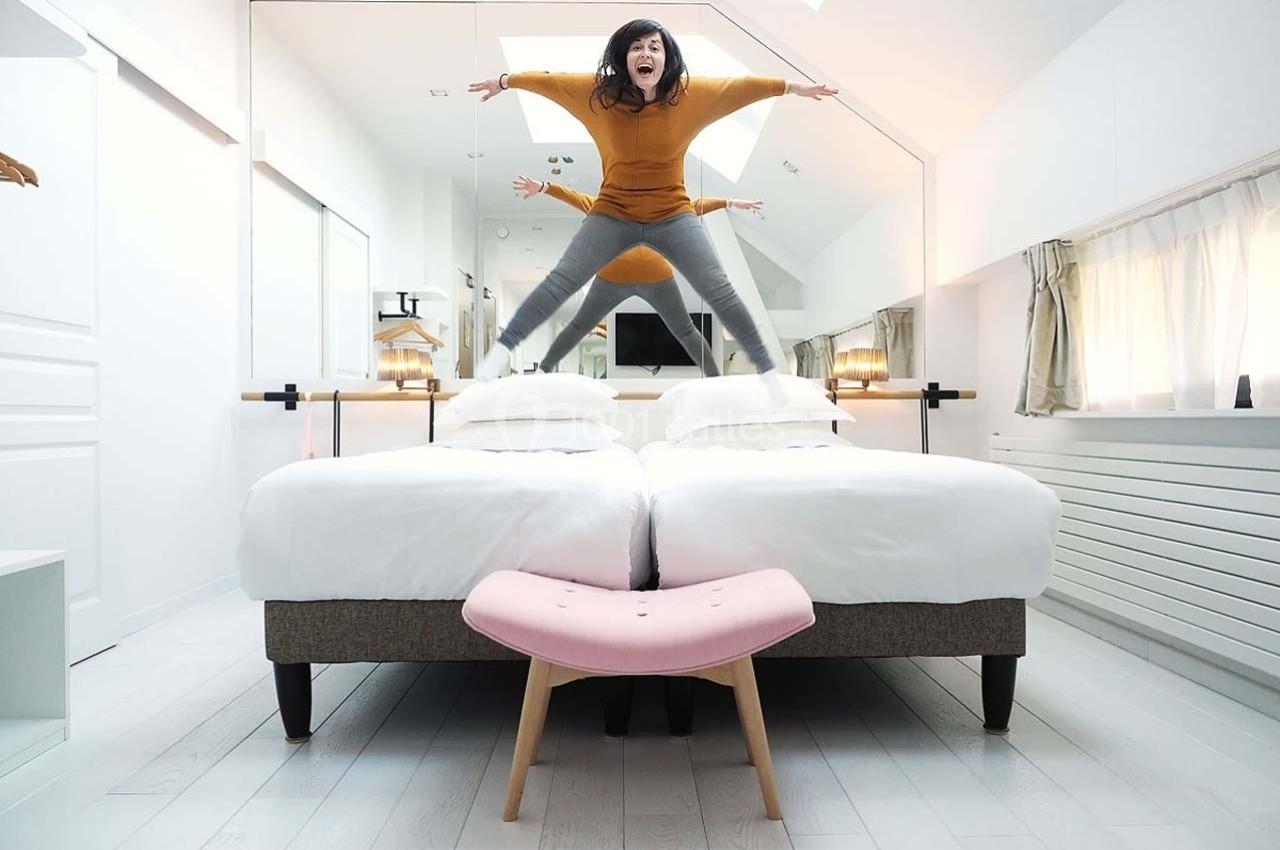 Une femme saute sur deux lits jumeaux dans une chambre lumineuse avec un miroir et un tabouret rose au premier plan.