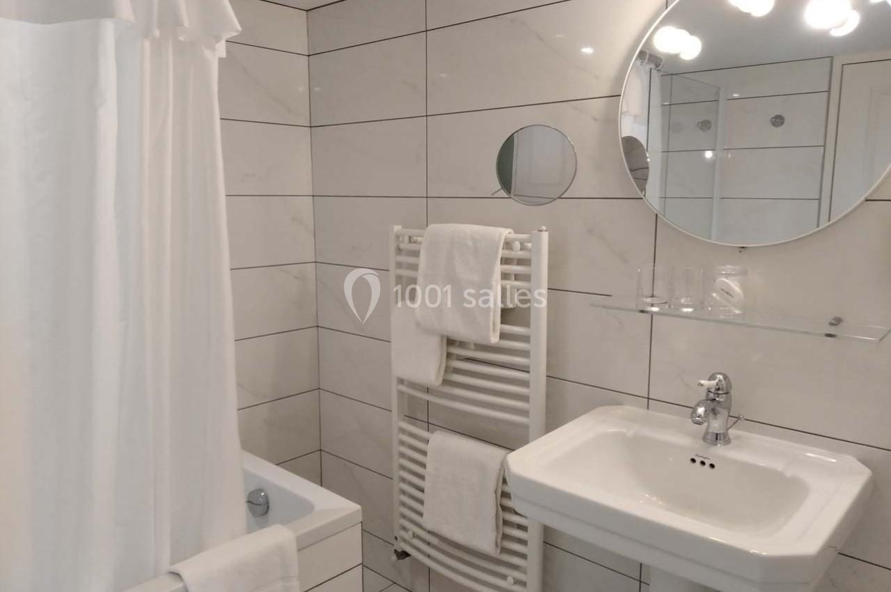 Salle de bain moderne avec carrelage blanc, lavabo, miroir rond, baignoire avec rideau et porte-serviettes chauffant.