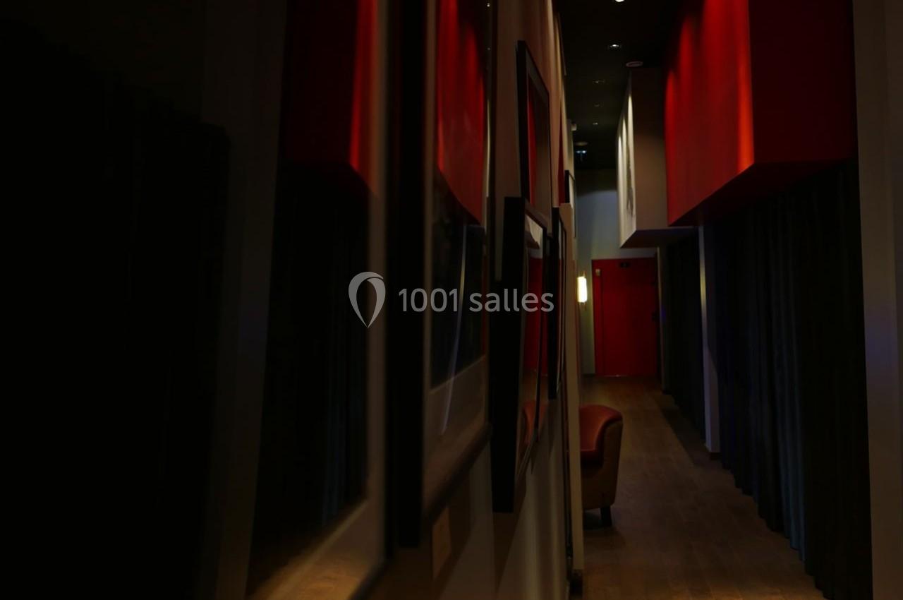 Couloir sombre avec murs rouges, éclairage tamisé et fauteuil visible au fond près d'une porte.