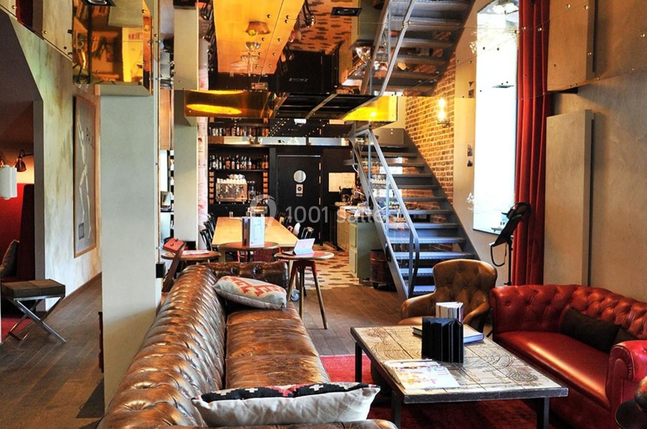 Salon moderne avec canapé en cuir, fauteuils rouges, table en bois, escalier métallique et éclairage chaleureux.