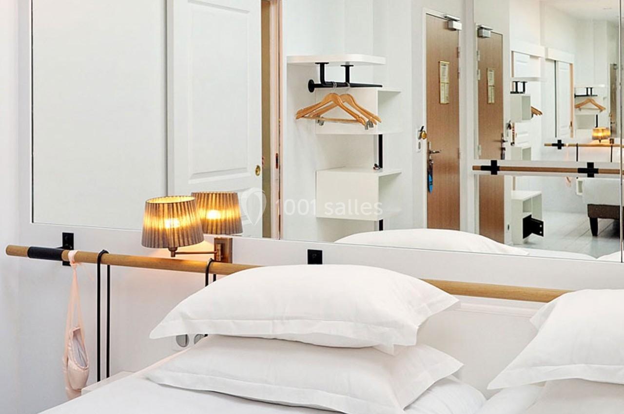 Lit avec oreillers blancs, lampe de chevet allumée et miroir reflétant une penderie et des portes dans une chambre lumineuse.