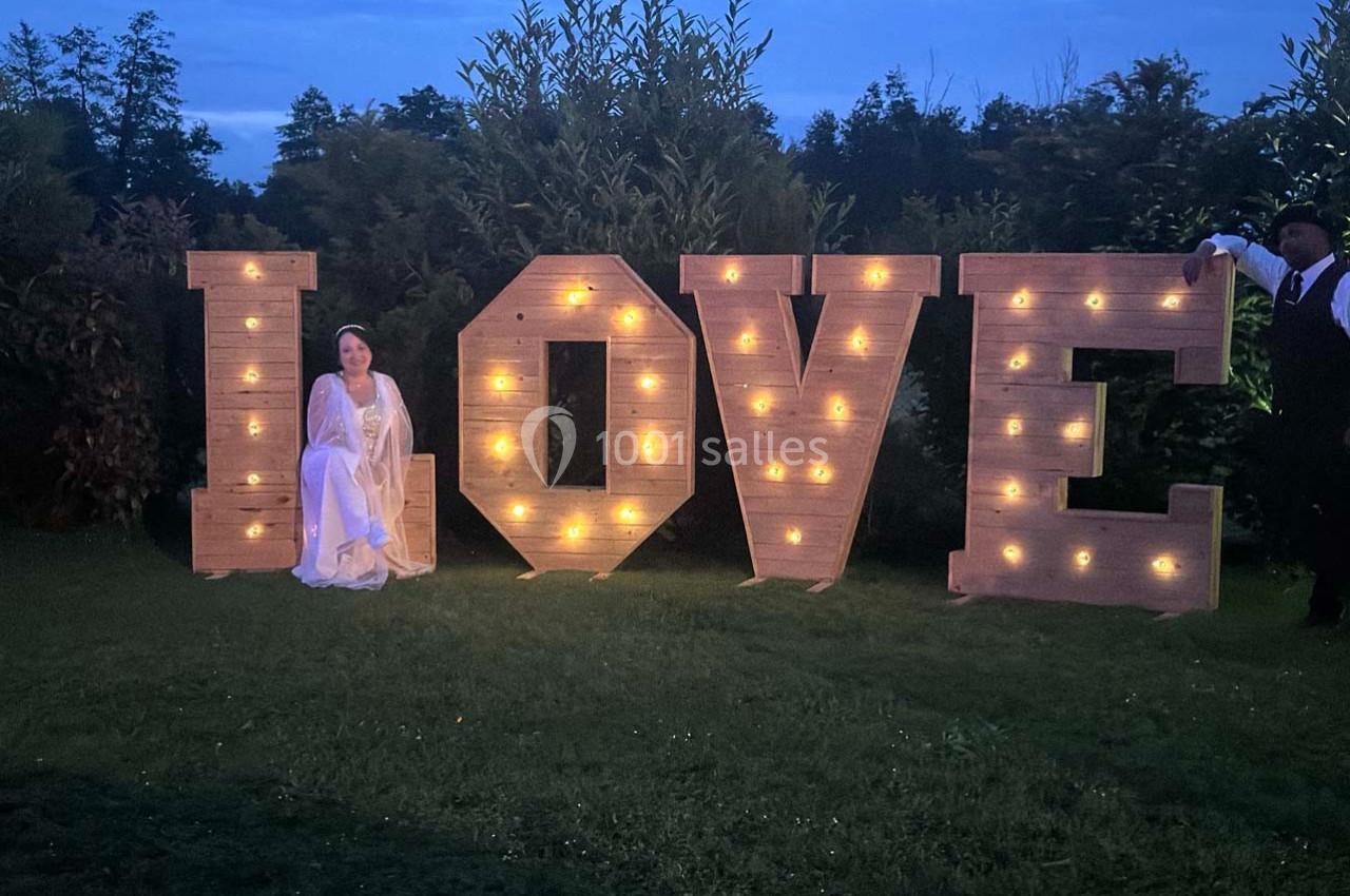Une femme en robe blanche et un homme en costume posent près de grandes lettres lumineuses formant le mot ’LOVE’ dans un…