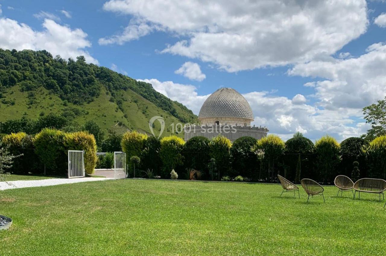 Pelouse verdoyante avec des chaises en osier, un bâtiment à dôme en arrière-plan et des collines boisées sous un ciel…