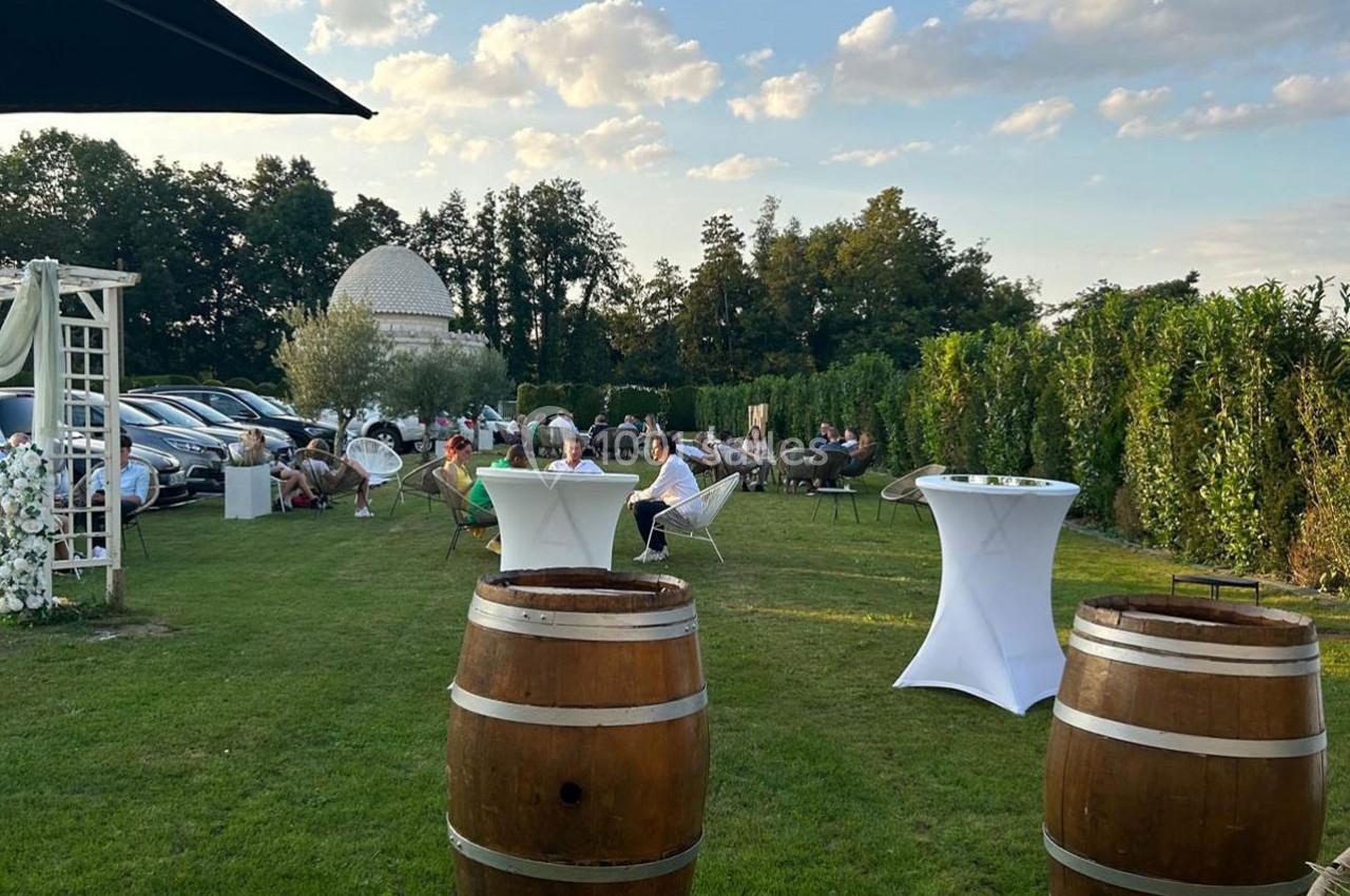 Personnes assises et debout dans un jardin avec des tables hautes, des tonneaux et une coupole blanche en arrière-plan.