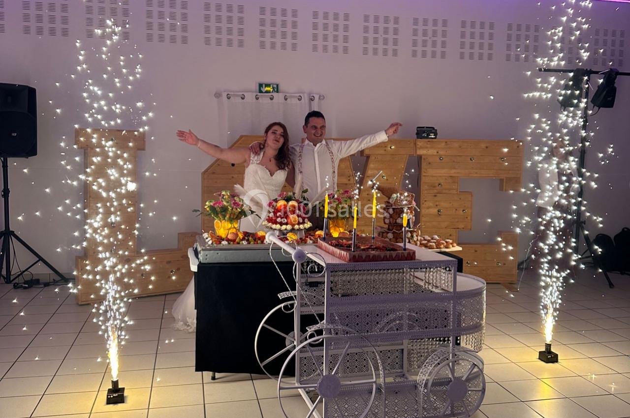 Un couple de mariés souriant pose devant une table décorée de desserts, entourée de fontaines lumineuses.