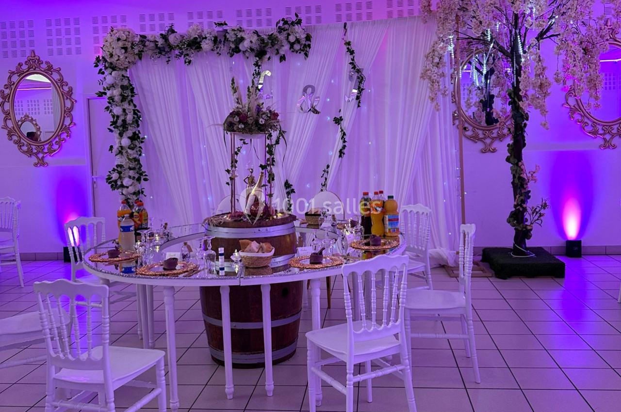 Table décorée pour un événement, entourée de chaises blanches, avec boissons, nourriture et décor floral en arrière-plan.