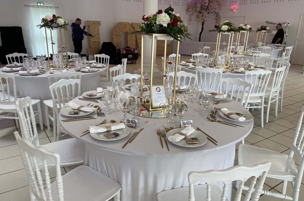 Salle de réception décorée avec des tables rondes élégantes, nappes blanches, centres de table floraux et couverts dorés.