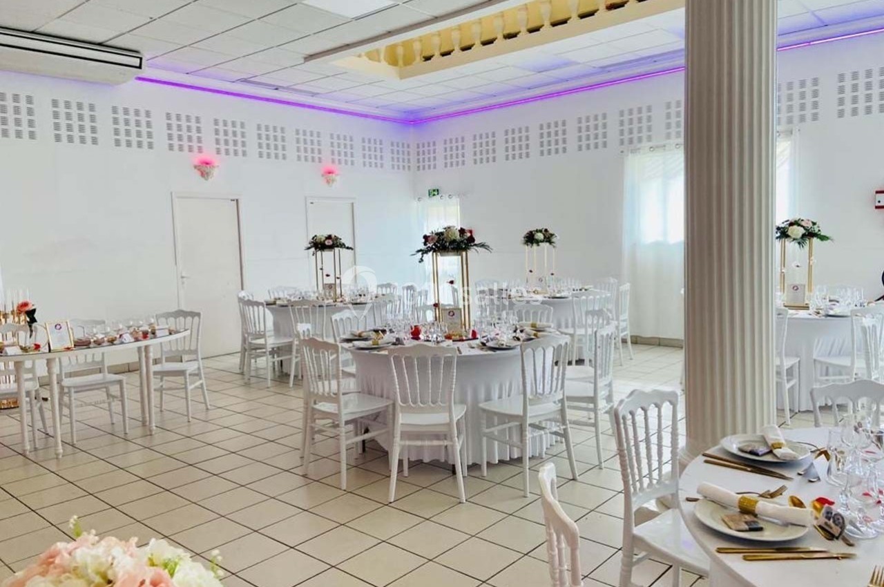 Salle de réception lumineuse avec tables rondes décorées de fleurs, chaises blanches et éclairage LED violet.