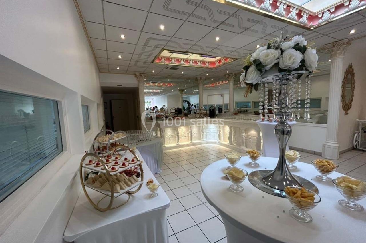 Salle de réception lumineuse avec décoration florale, buffet de desserts et tables dressées en arrière-plan.