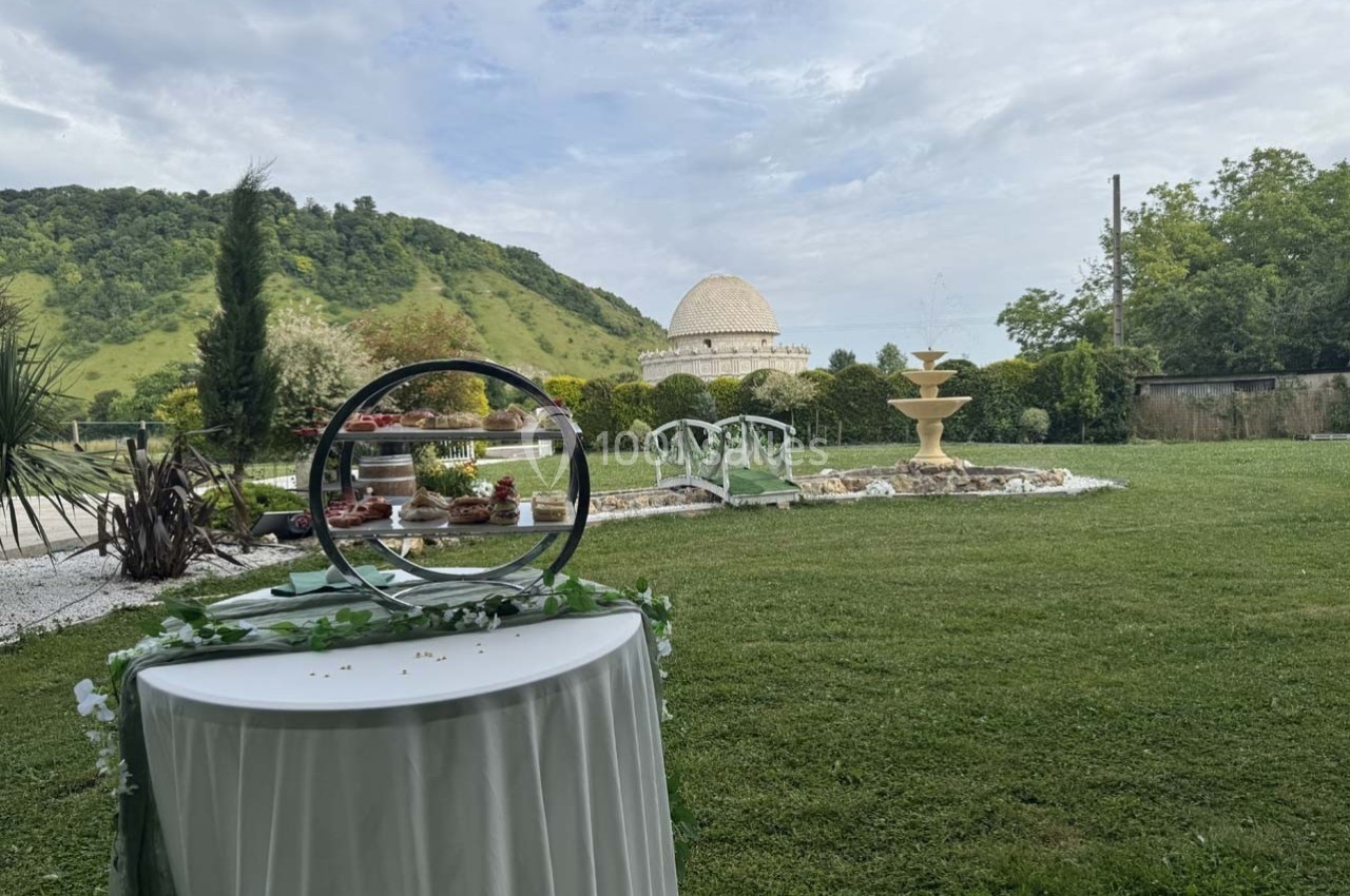Jardin verdoyant avec une table décorée au premier plan, une fontaine et un bâtiment rond en arrière-plan.