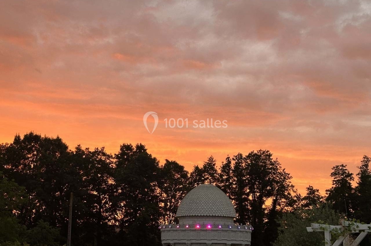 Coupole blanche éclairée au crépuscule, entourée d'arbres sombres sous un ciel orangé nuageux.