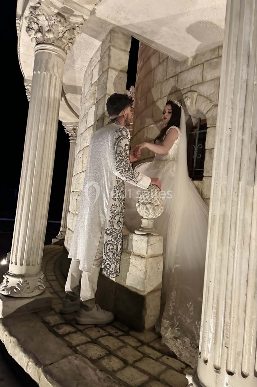 Un couple en tenue de cérémonie se tient sous une structure en pierre avec des colonnes, éclairée de nuit.